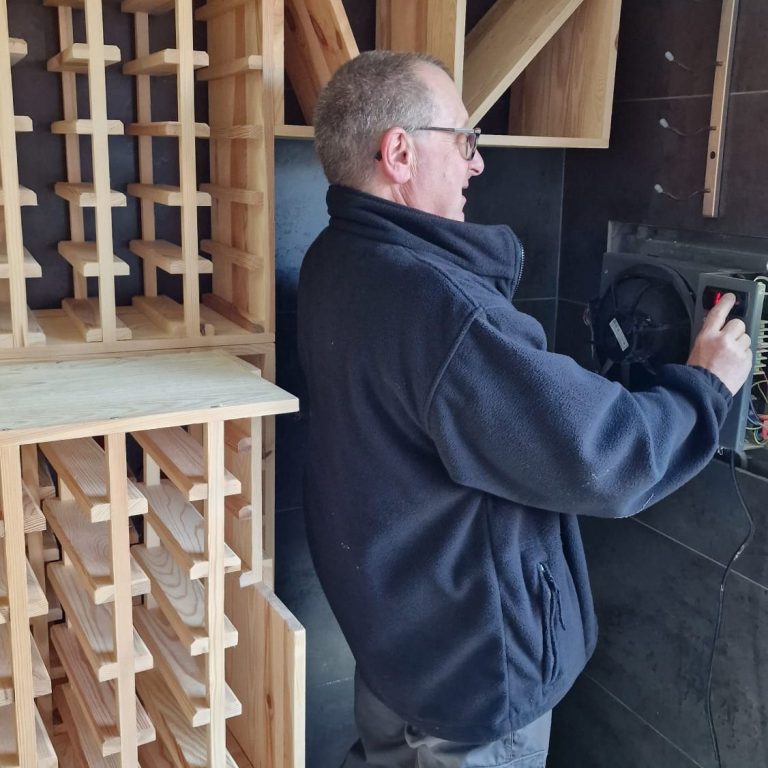 Steve M. A man adjusts equipment in a wooden storage area with wine racks.